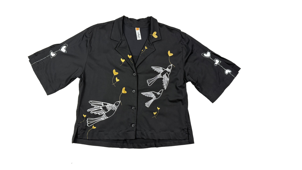Aves y corazones en delicadas líneas doradas y blancas: el arte gráfico que transforma una camisa básica en una pieza de autor.