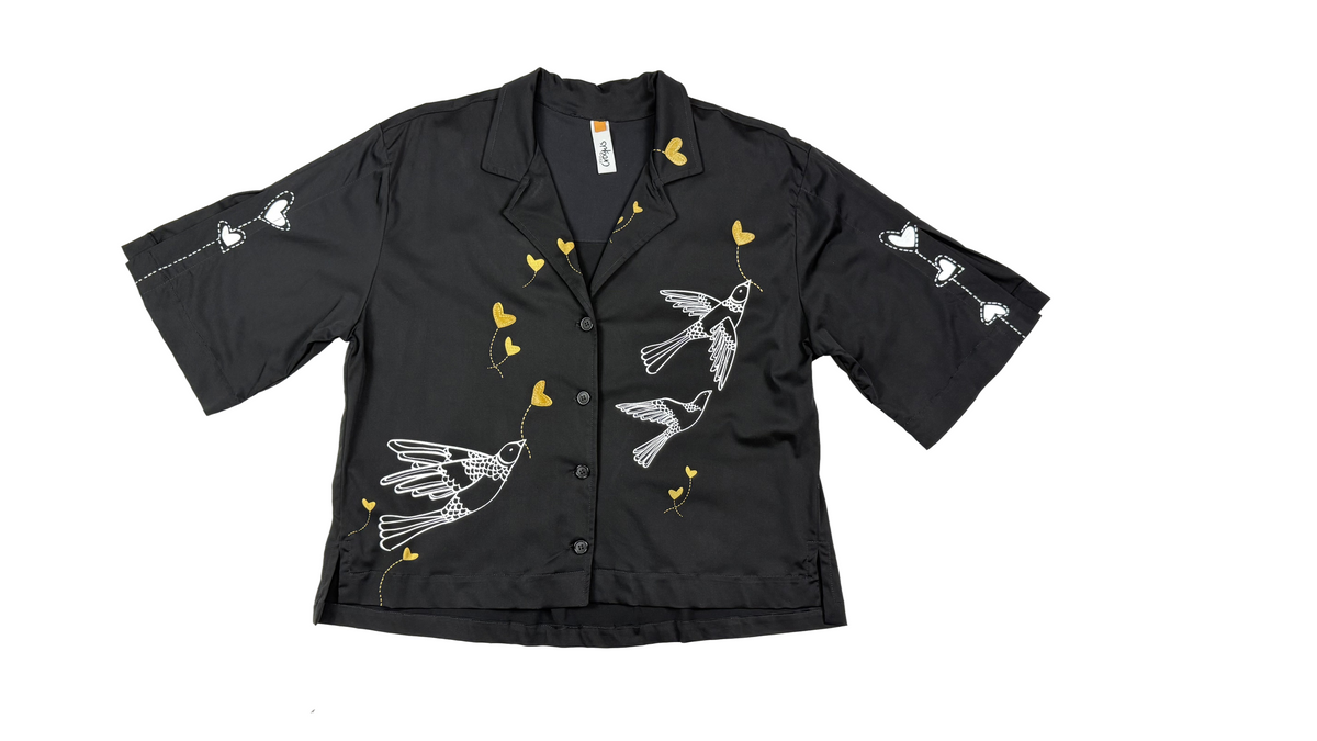 Aves y corazones en delicadas líneas doradas y blancas: el arte gráfico que transforma una camisa básica en una pieza de autor.